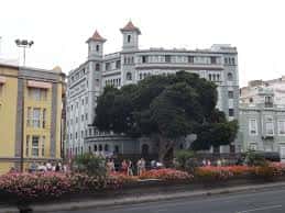 Fachada del teatro desde la plaza, con árboles y flores en primer plano