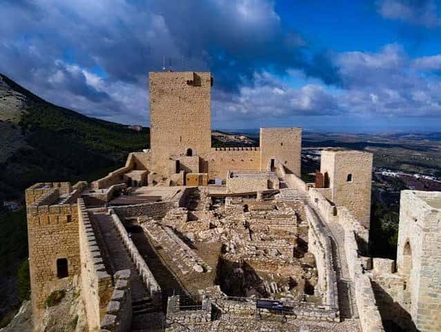 Plaza interior del castillo con murallas, almenas y vistas al valle bajo nubes dramáticas