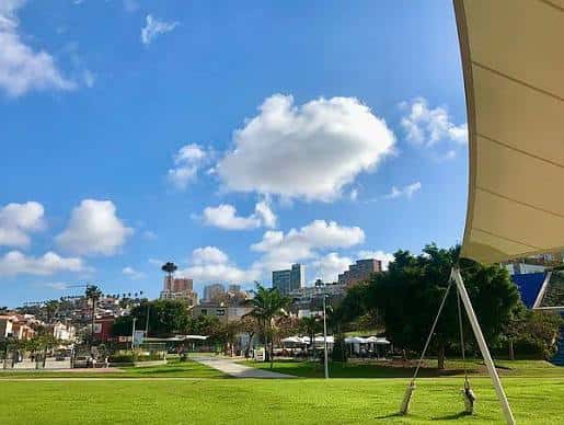 Vista panorámica del parque con césped, árboles y la estructura del estadio al fondo bajo un cielo azul