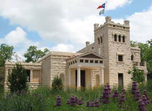 Fachada principal del edificio histórico rodeado de flores púrpuras bajo cielo azul