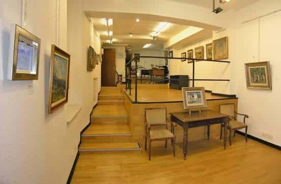 Interior de la Galería Cervantes mostrando las salas de exposición con obras enmarcadas y escalera central