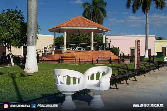 Paseo ajardinado frente a la ermita con bancas blancas y kiosco