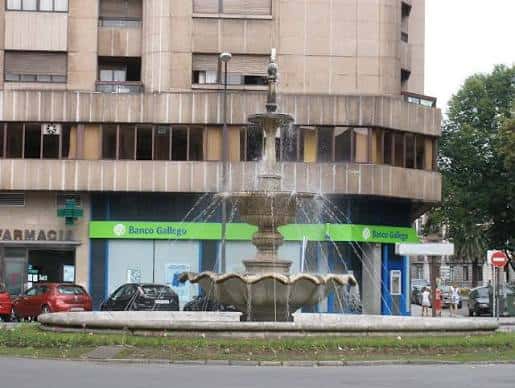 Vista de la plaza con fuente en primer plano y edificio moderno con farmacia y banco