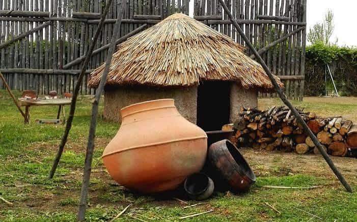 Reconstrucción de una cabaña prehistórica con techo de paja y grandes vasijas cerámicas en primer plano