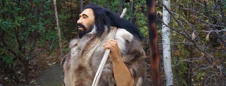 Figura de un hombre de las cavernas con vestimenta de pieles y bastón en entorno forestal