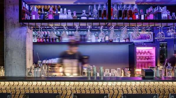 Interior del bar con estantería de botellas, copas colgantes y barra iluminada