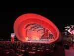 Vista nocturna del escenario iluminado en rojo con el público sentado frente a él