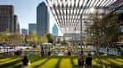 Plaza soleada de Sammons Park con césped, sombra de la estructura y rascacielos al fondo