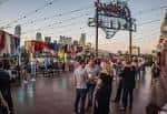 Plaza exterior de Gilley's Dallas al atardecer con gente caminando bajo luces colgantes y vista del skyline de Dallas