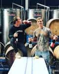 Dos personas disfrutando de una cata de vinos en la bodega con tanques de acero y barricas al fondo