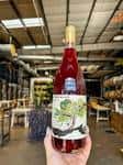 Mano sosteniendo una botella de vino rosado frente a la bodega con barricas y mesas de madera
