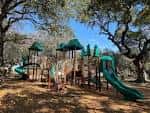 Área de juegos infantil con toboganes verdes y estructuras de madera bajo árboles frondosos