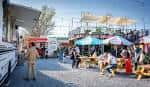 Área de eventos al aire libre con toldos, puestos de comida y público en un entorno urbano