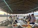 Vista interior de la cubierta del barco con techo de paja y personas disfrutando de una comida al atardecer