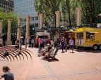 Escena animada en la plaza con personas caminando, food trucks amarillos y bancos curvos bajo árboles frondosos