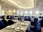 Salón preparado para banquete con mesas largas, sillas azules y vajilla dispuesta