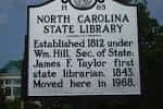 Letrero histórico de la North Carolina State Library con detalles arquitectónicos al fondo