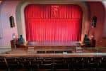 Vista frontal del escenario del teatro con butacas vacías y cortina roja cerrada