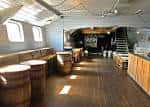 Interior del Grain Barge con barra de madera, barriles decorativos y escalera hacia la cubierta