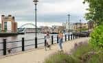 Paseo junto al río con personas caminando y el Puente del Milenio al fondo