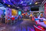 Interior del bar con iluminación ambiental colorida, mesas altas y zona de música en vivo
