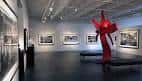 Sala de exposición con escultura roja central y cuadros enmarcados