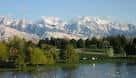 Lago rodeado de árboles y césped con vista a las montañas en primer plano