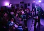 Interior del club de comedia con público y comediante en el escenario bajo luces púrpuras