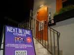 Entrada nocturna del Next In Line Comedy Club con letrero iluminado y escalera de acceso