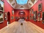 Interior de una sala de exposiciones con paredes rojas, cuadros colgados y techo con cúpula de cristal