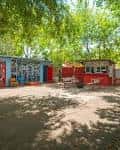 Área exterior diurna con contenedores pintados y mesas bajo árboles