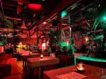 Interior del lounge con mesas y decoración tropical bajo luces rojas y verdes