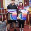 Dos personas sonriendo mientras sostienen sus pinturas terminadas en un taller de Painting with a Twist