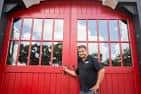 Fachada principal con grandes puertas rojas y un hombre posando junto a ellas