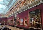 Interior de una sala de exposiciones con cuadros colgados en paredes rojas y techos ornamentados