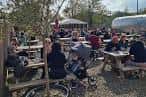 Área al aire libre de Redwell Brewery con personas disfrutando en mesas bajo árboles