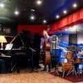Escenario interior con músicos tocando piano, contrabajo y batería bajo luces rojas