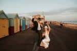 Pareja celebrando en la playa frente a los coloridos chiringuitos de Brighton, mostrando el entorno del lugar