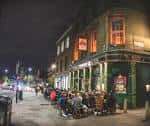 Vista nocturna de la fachada exterior del pub con clientes sentados en la acera