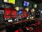 Área del bar con estantería de botellas iluminadas en rojo y múltiples pantallas colgantes