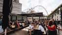 Interior al aire libre con mesas, personas comiendo y estructura de contenedores bajo un puente