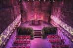 Salón con paredes de piedra y escenario iluminado en tonos púrpuras, ideal para ceremonias o recepciones