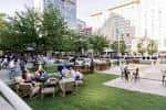 Área de descanso con mesas y sillas en Gallivan Center rodeada de árboles y edificios modernos