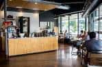 Interior del café con mostrador de madera, clientes sentados y ventanas luminosas