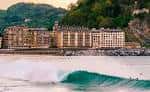 Primer plano de surfistas en una ola con los edificios de la Zurriola al fondo