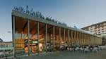 Vista exterior del moderno Mercado de Abastos con fachada de cristal y estructura de madera, iluminado al atardecer