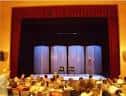 Interior del salón de actos o teatro con escenario y cortinas rojas, y público asistente