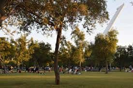 Pradera arbolada con personas realizando actividades al aire libre y el puente del Alamillo al fondo