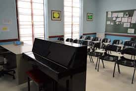 Interior de un aula de música con piano vertical, pupitres y pizarra