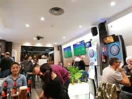 Interior del local con mesas, pantallas de televisión y zona de juegos con diana